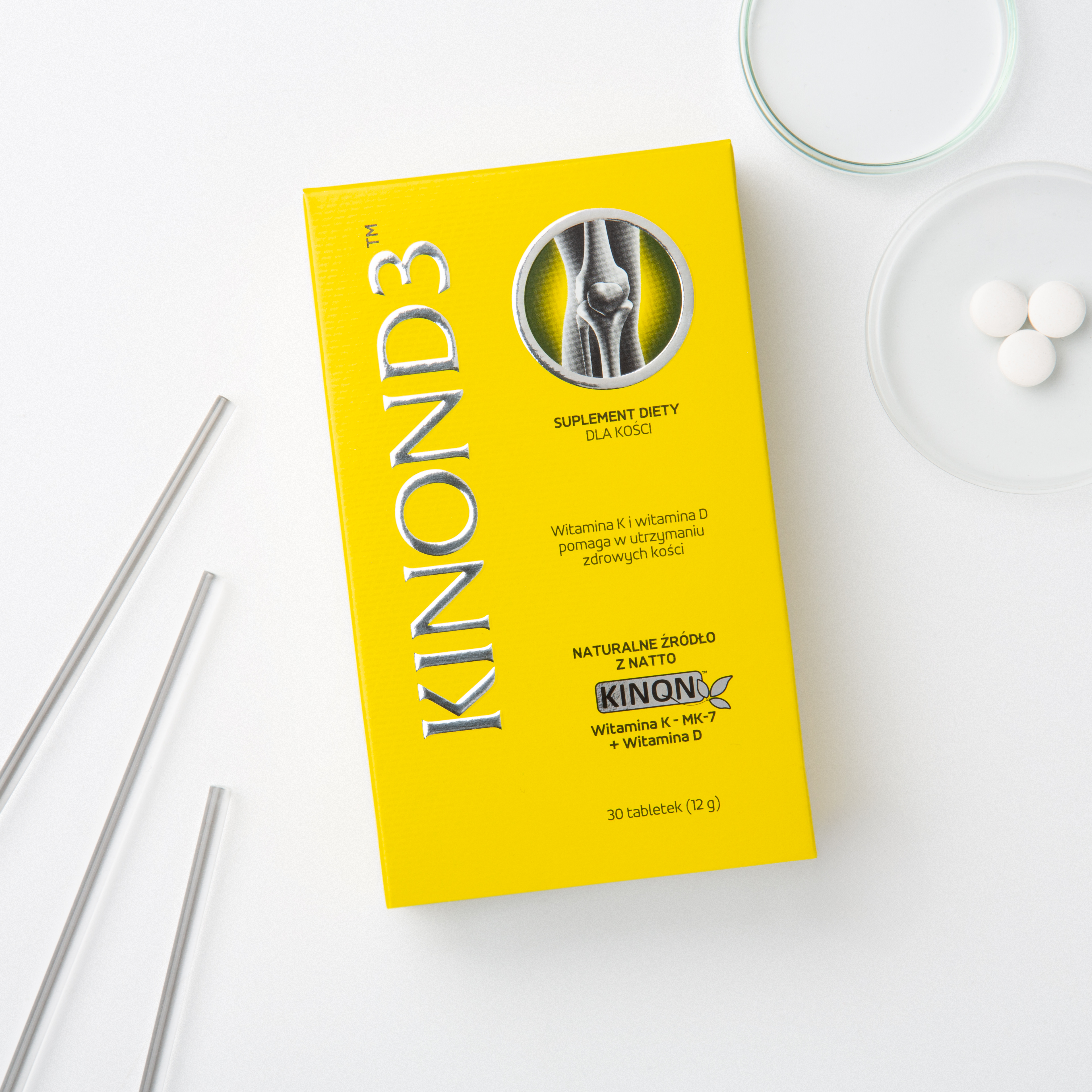 Intensywnie żółte opakowanie suplementu Kinon D3 na jasnym tle. Obok pudełka, na przezroczystej szalce, leżą trzy małe, białe tabletki. Na opakowaniu widoczny jest srebrny napis KINON D3, grafika stawu kolanowego oraz informacja o naturalnym źródle z Natto.
